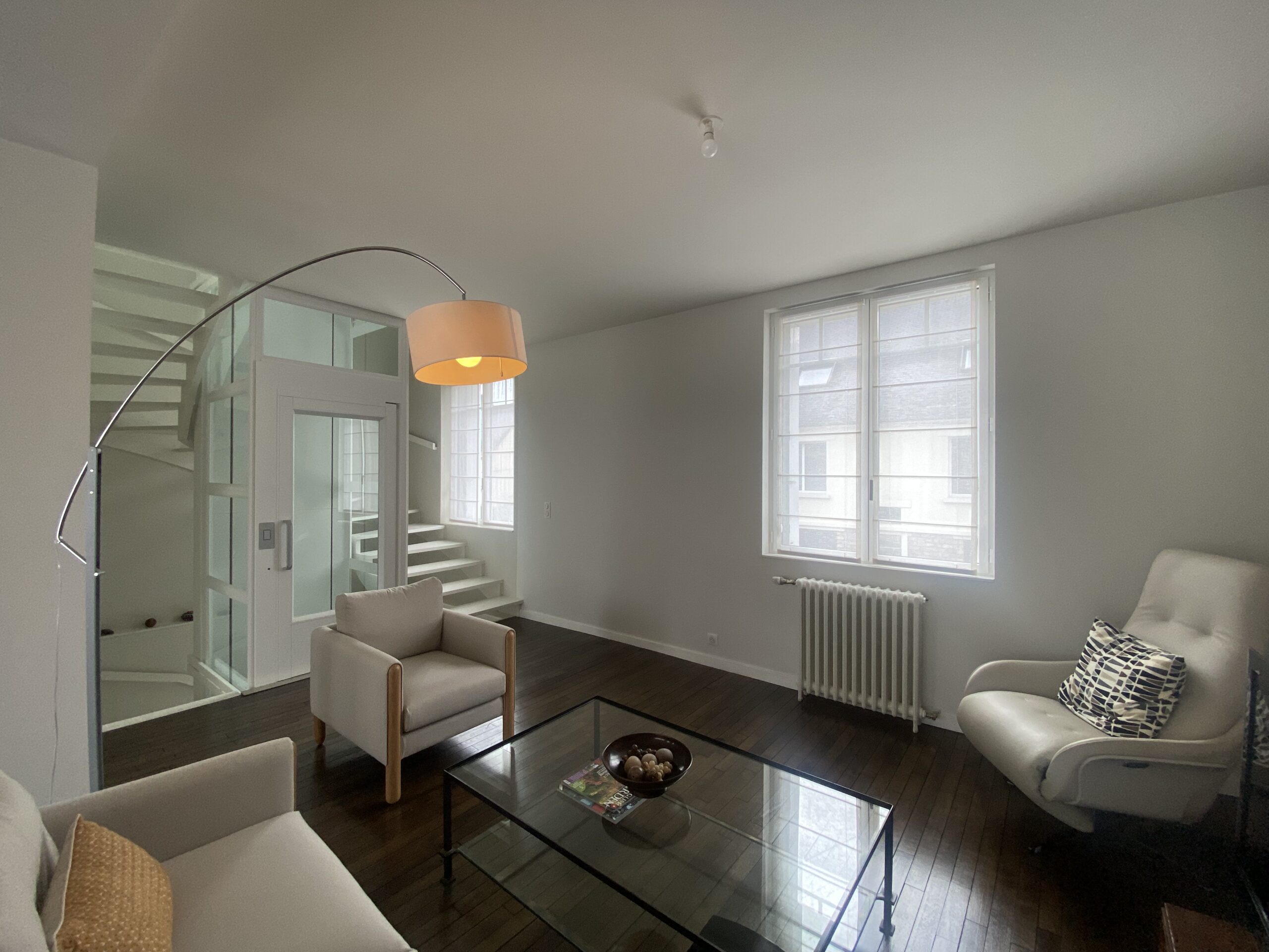 Salon design contemporain avec fauteuils clairs, lampadaire arc et escalier architectural avec monte charge.